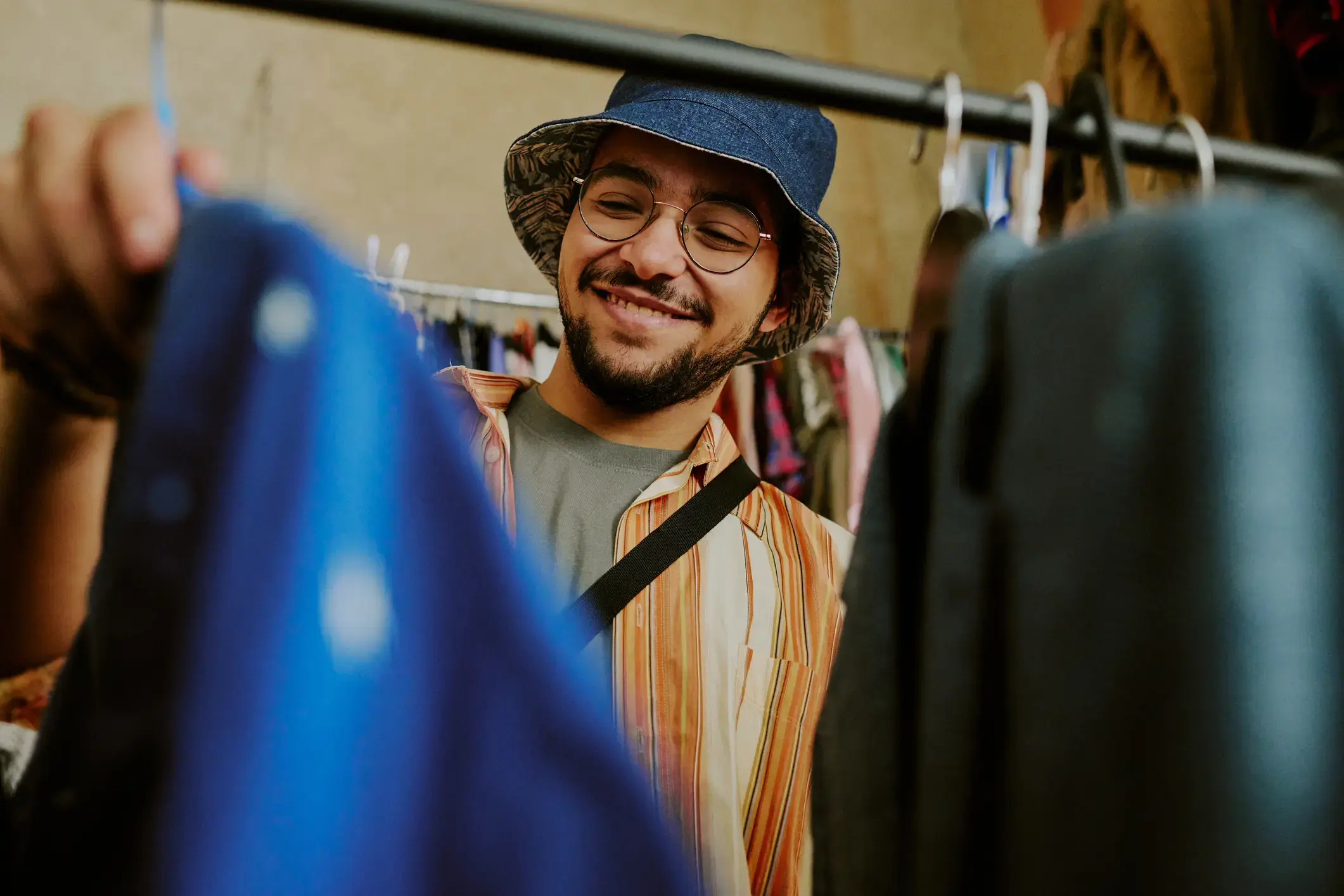 Young man wearing glasses with casual clothing and hat browsing clothes in thrift store while smiling cheerfully, engaged in the joy of shopping for unique apparel.